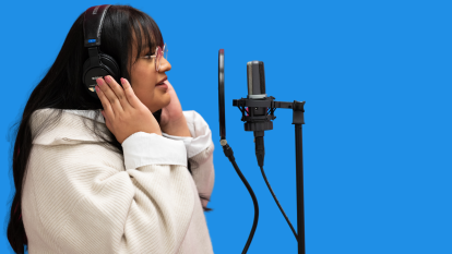 A woman with dark hair holds her hands over headphones while speaking into a microphone in front of a blue background.