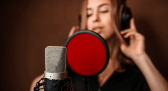 A woman wearing headphones stands behind a microphone with a pop filter attached to it.